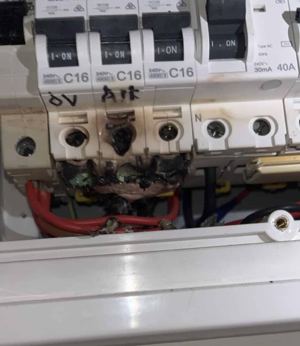 Exposed electrical circuit breaker with burnt marks, mounted inside a white electrical panel on a wall, showing signs of damage and melting.