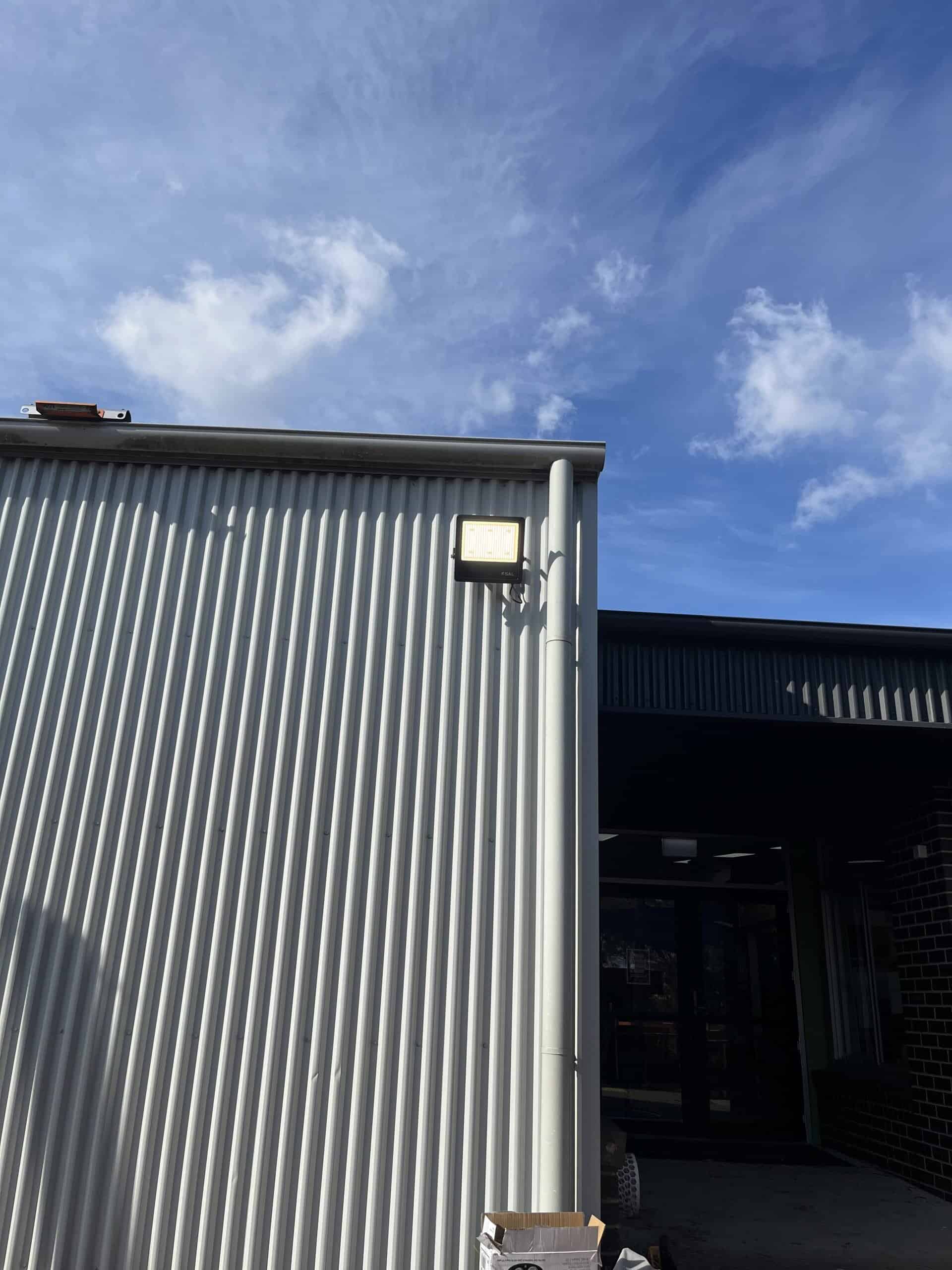 Outdoor wall-mounted LED floodlight on a corrugated metal surface in a commercial or industrial setting, with a partly cloudy sky overhead.