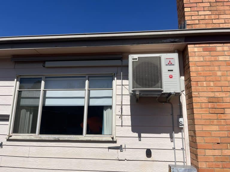 External air conditioning unit mounted on the wall of a house, underneath a window, outside in daylight.