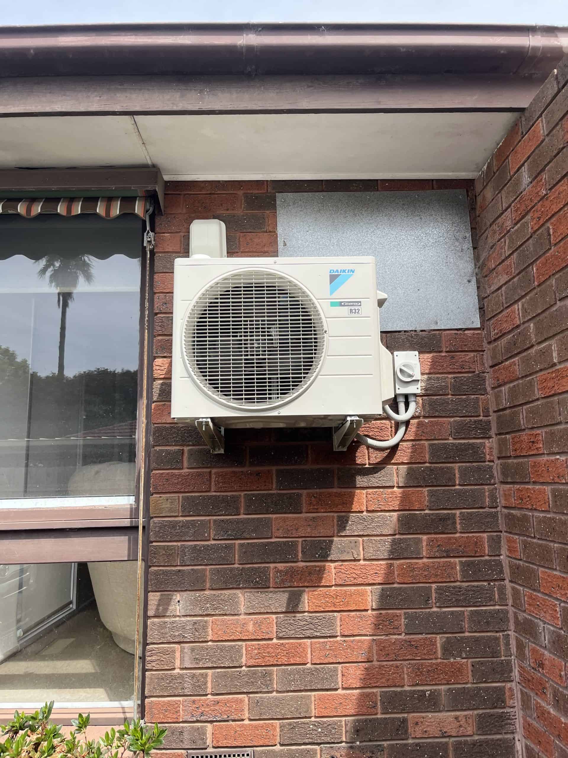 Performing a maintenance check on the outdoor air conditioning unit mounted on a brick wall. The unit is installed on a concrete surface beneath a roof overhang.