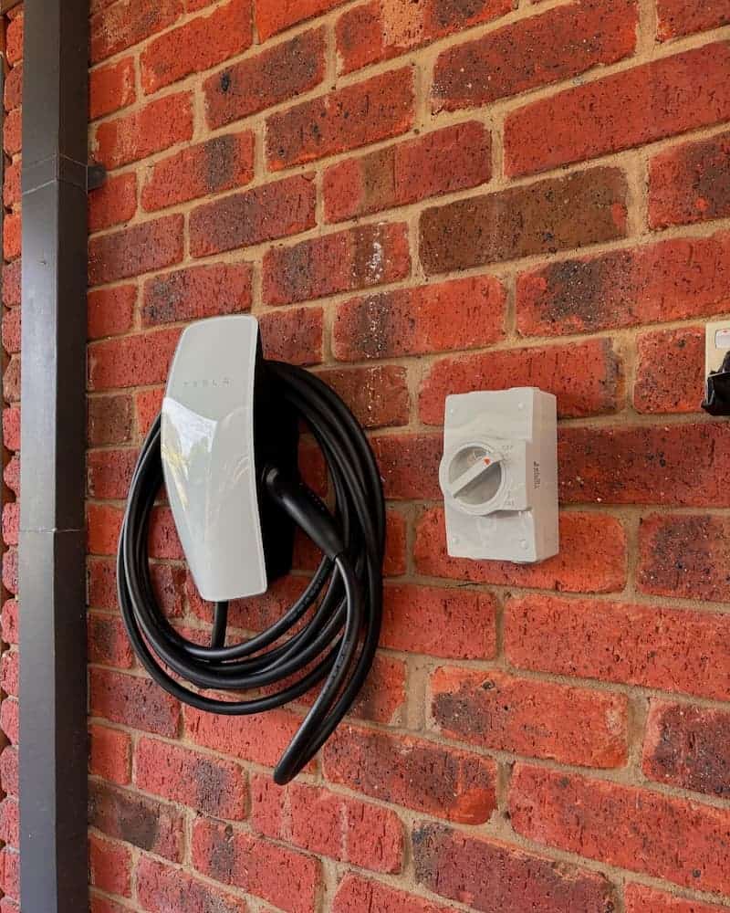 EV charging station mounted on a red brick wall outside a building, with a coiled black cable and white plug protector.