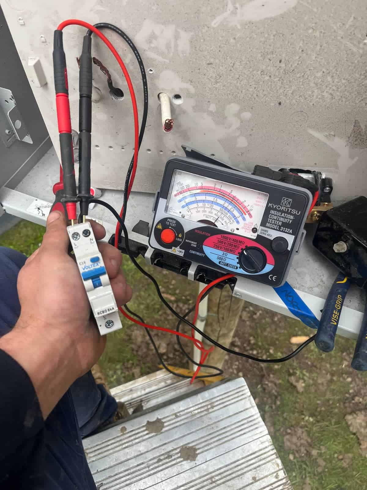 Clamp meter testing electrical wiring on an outdoor metal electrical box surface.