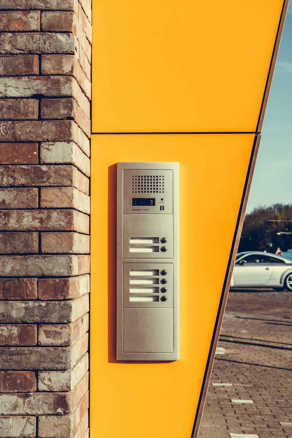 Intercom system mounted on a yellow exterior wall beside a brick wall outside building.