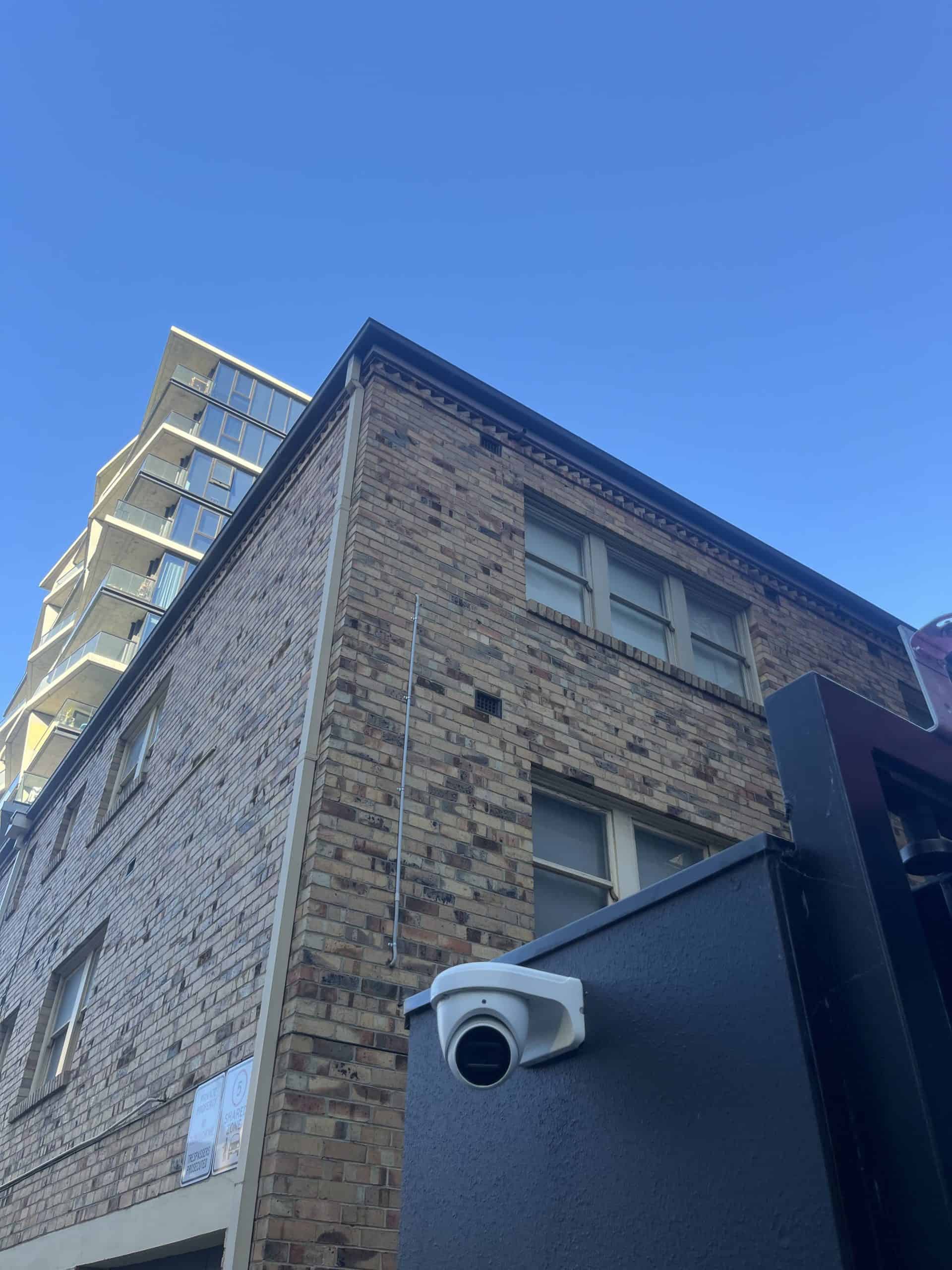 CCTV security camera mounted on a dark painted brick exterior wall of a building, situated outdoors under a clear blue sky.