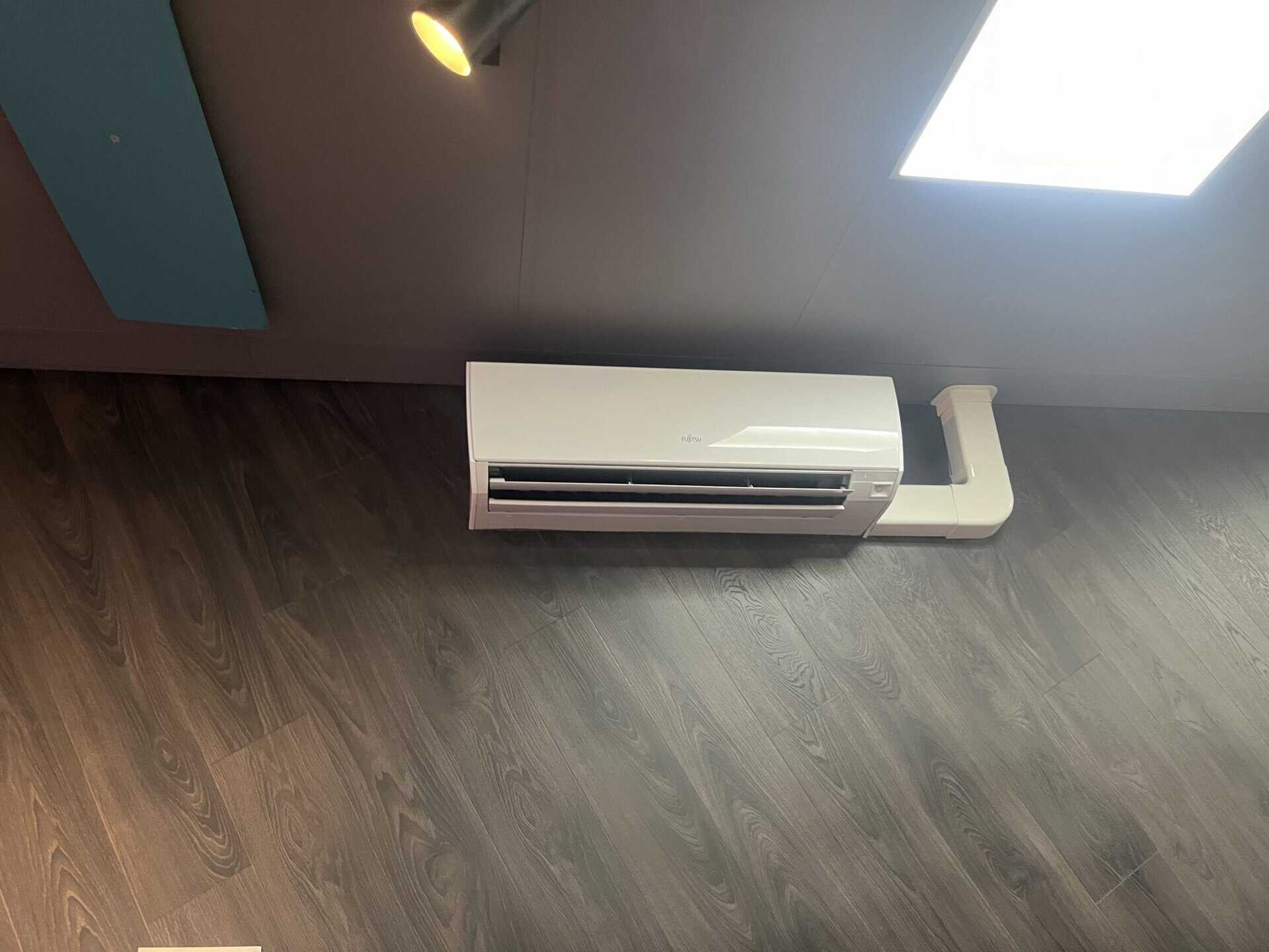 Air conditioning unit mounted on an indoor wall with a dark ceiling and wooden floor surface.