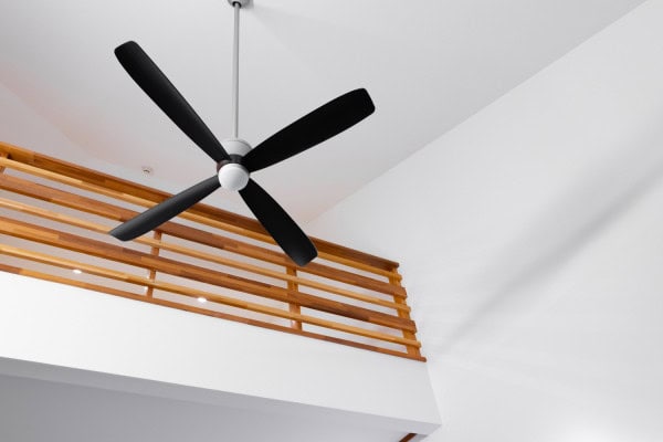 Black ceiling fan with four blades mounted on a white indoor ceiling near a wooden railing, inside a modern building.