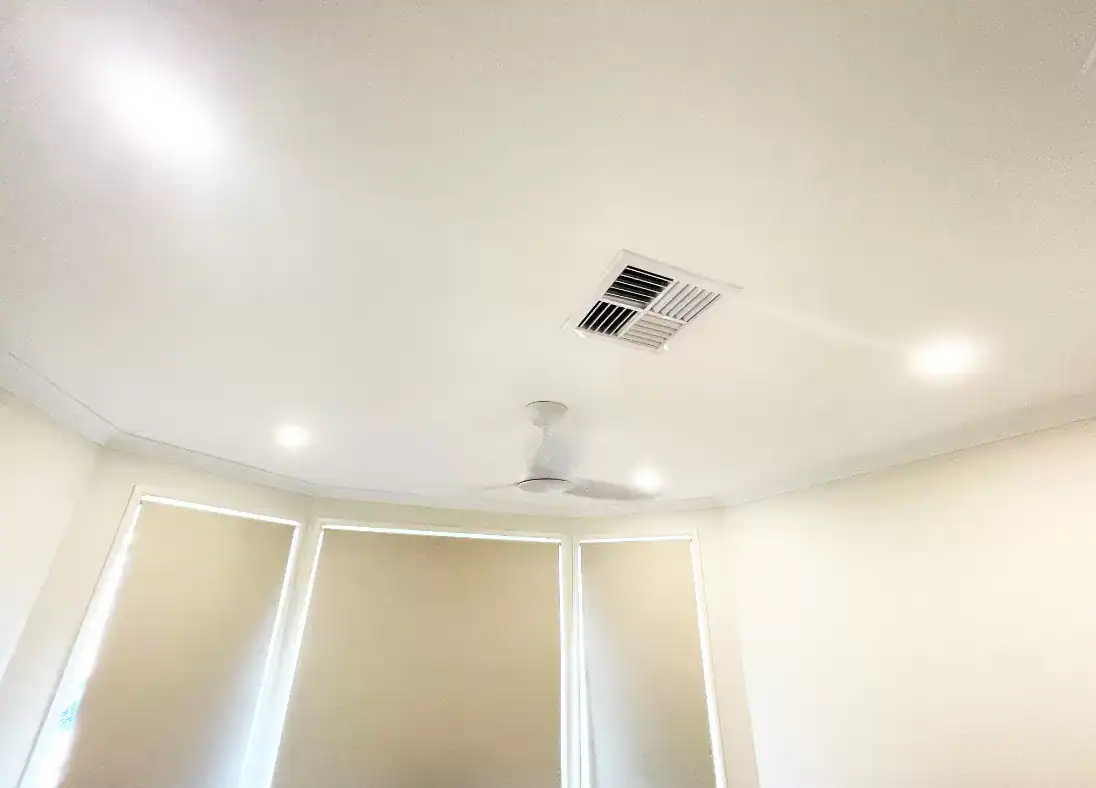 Modern indoor ceiling with air conditioning vent and ceiling fan on a smooth white surface.