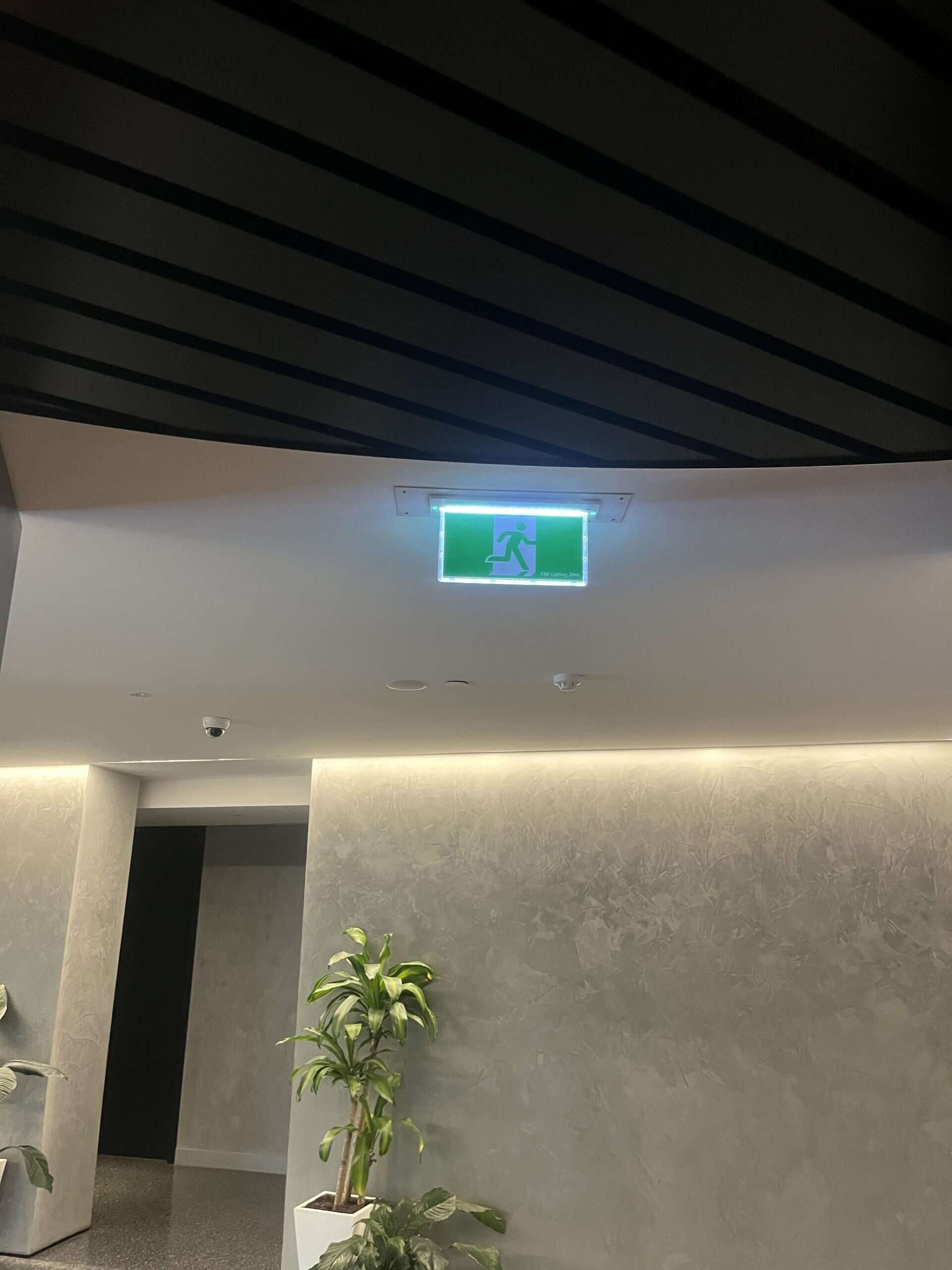 Emergency exit sign with green background and running figure, mounted on a white ceiling inside a building, illuminated for visibility.