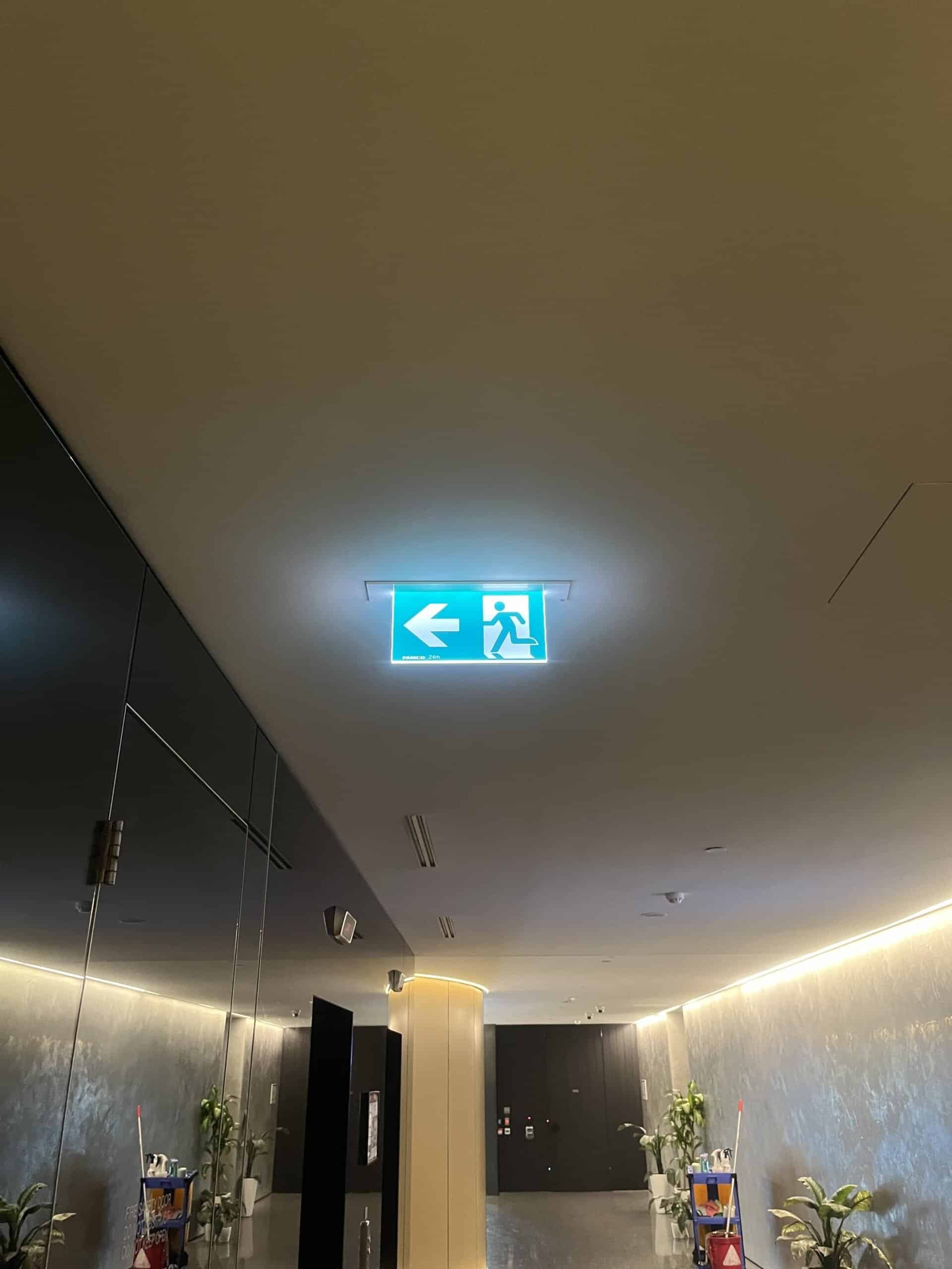 Illuminated emergency exit sign on ceiling indoors in a modern building.