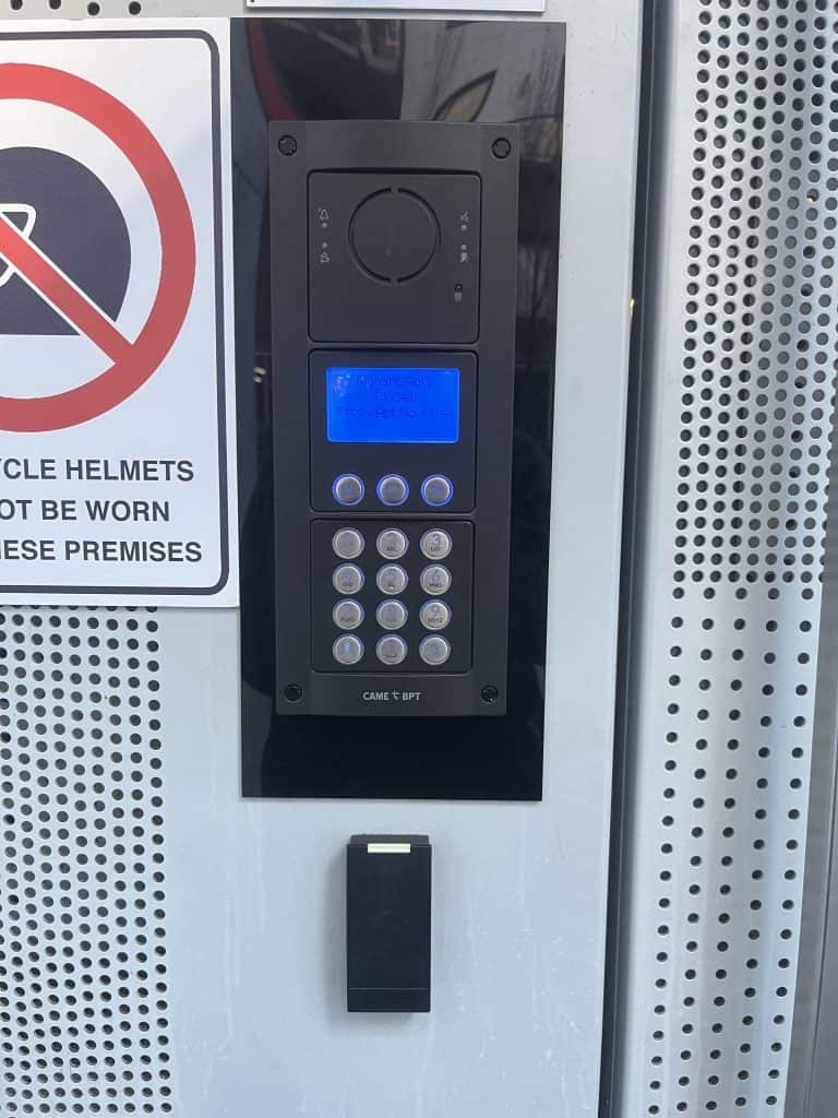 Alarm panel with a keypad and blue digital display mounted on an outdoor metal surface with perforated pattern.