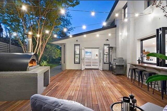 Outdoor wooden deck with string lights, open wall window, and outdoor pizza oven on grey concrete base.