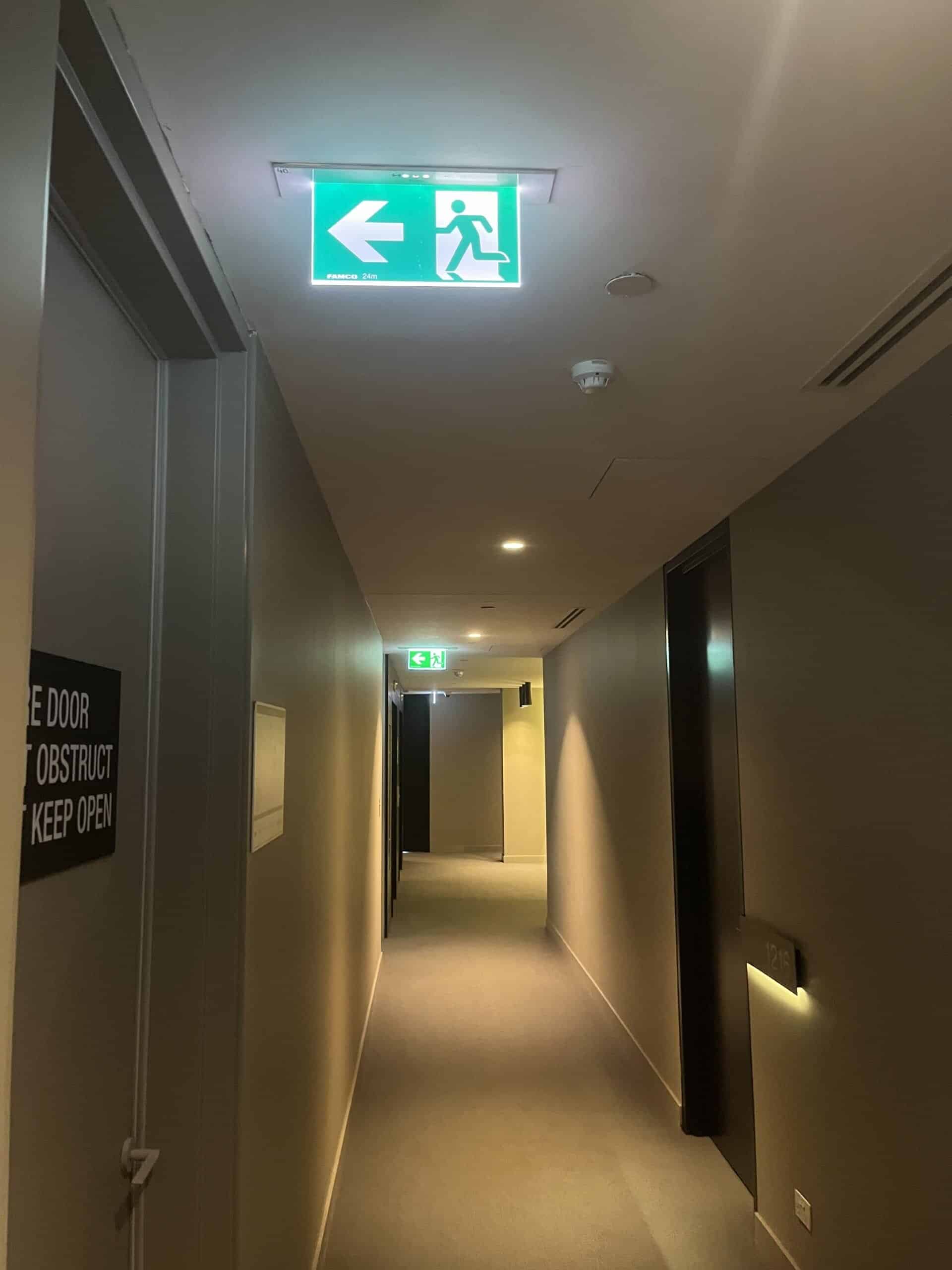 Exit sign with green background and white symbols, mounted on indoor ceiling of hallway.